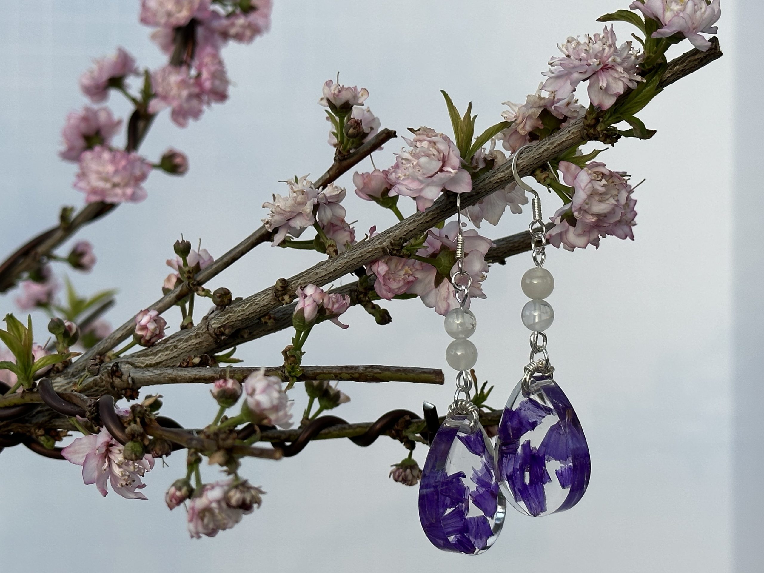 Earrings hanging on a bonsai tree with pink flowers. The earrings are sterling silver with moonstone beads and teardrop resin pendants with dried wildflower inclusions.